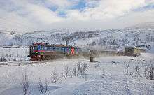 A three-unit locomotive hauls an ore train out of a tunnel, surrounded by snow.