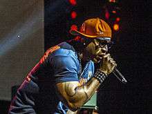 On a brightly-lit stage, an African-American man wearing an orange baseball cap, glasses and a blue shirt raps into a microphone.