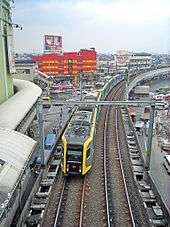Below, on elevated tracks surrounded by a daytime urban landscape of roads, cars, buildings, and billboards, a metro train approaches, its front driver's window framed on the sides by angled planes and yellow markings visible as the rest of the train trails behind curving slightly to the right into the distance.