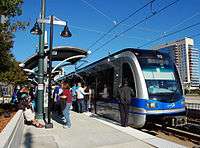 A blue and gray train stopped at a covered, side platformed station with several passengers entering.