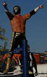 The wrestler La Sombra standing on the second rope of a wrestling ropes, pointing to the fans during an outdoor event.
