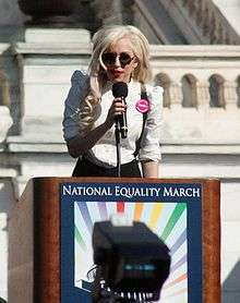 A blond woman wearing a white shirt and black glasses speaking on a lectern carrying a 'National Equality March' poster. Behind her is a white stone balustrade of a building.
