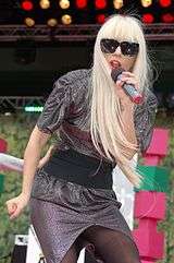 Lady Gaga with long blond hair, wearing a grey dress, a black belt and black sunglasses, singing into a microphone on an outdoor stage