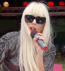A woman with long blond hair and black sunglasses holding a microphone up to her mouth