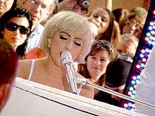 A blond woman with short cropped hair, singing to a microphone, in front of a piano. Faces of the crowd can be seen behind her.