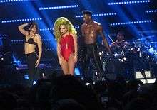 In the center of the photograph is a woman with blonde hair in red clothing; to her sides are two scantily-clad people, and in the background is someone sitting at a drum set.