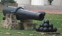A long 19th-century cannon with a stack of cannon balls in front of it, sitting on a lush green lawn.