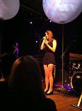 A brunette female sings on a stage in front of a crowd, wearing a black dress that covers above the knees and black high heels. A blue balloon obscures the upper righthand corner.