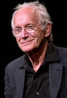 The image is of an older man with glasses wearing a black shirt. He is looking past the camera, smirking.