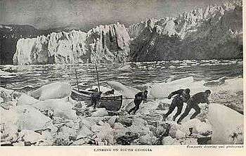  Six men pulling a boat on to an icy shore, with a line of ice cliffs in the background