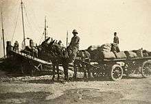 Two mules pulling a wagon loaded by supplies. A man rides one of the mules, while another man stands on the wagon.