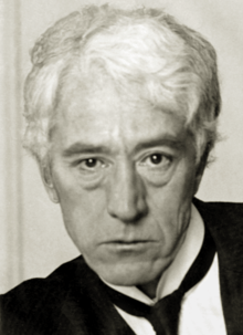 A white-haired man in his fifties, wearing a black jacket and tie and white shirt