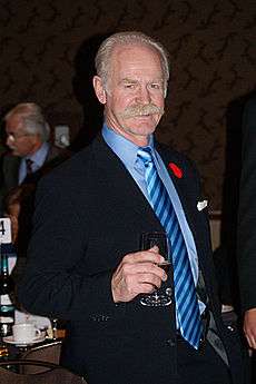 A grey-haired, grey-moustached man holds a wine glass.  He is wearing a blue suit with a striped blue tie