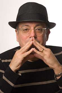Picture of a man in his 50s. He wears glasses and a fedora hat. He holds his both hands in front of his mouth.