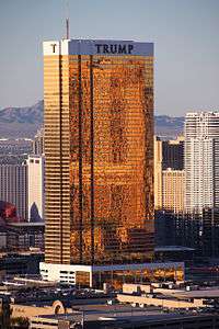 A tall rectangular-shaped tower in Las Vegas with exterior windows shimmering with 24-karat gold. It is a sunny day and the building is higher than many of the surrounding buildings, which are also towers. There are mountains in the background. This tower is called the Trump Hotel Las Vegas.