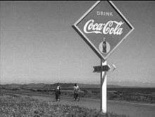 A young Japanese man and woman, both in casual clothes, are riding bicycles over a paved road in the near background of the image; mountains are visible in the far distance. In the foreground of the image, at the edge of the road, is a diamond-shaped Coca-Cola sign, below which is an arrow upon which is written the name (in English) of a beach.