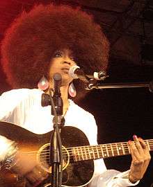An African-American female with a black afro strumming on a guitar in front of a microphone.