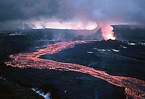 A red-hot lava flow streams out of a fuming vent, meandering past the viewer under a low cloudy sky.