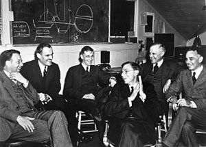 A group of in suits men sit around, laughing. On the blackboard behind is a discarded plan for assembling a uranium core.