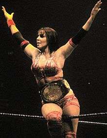 A dark-haired, tanned Caucasian female straddles the top rope of a wrestling ring with blue ropes. She is wearing a red patterned crop top, and matching shorts and kneepads. She is holding a wrestling championship above her head with both hands.