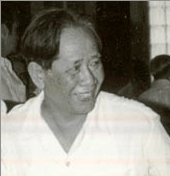A smiling man looking to the right, wearing a collarless white shirt