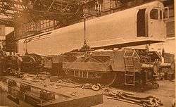 A picture of an 0-6-0+0-6-0 steam locomotive under construction. The cuboid box containing cabs and boiler is being lowered onto the two 0-6-0 bogies by a crane inside a locomotive works. Various components are scattered in the foreground.