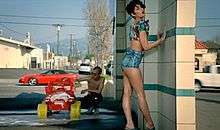 A woman wearing a crop top and short skirts poses in a carwash while a man washes a toy car in the background.