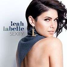 An image of a brunette woman looking over her shoulder. She wears an earring with a charm in the shape of an "L" and a denim top covering her neck and parts of her upper back. The words "leah labelle" and "SEXIFY" are written beside her.