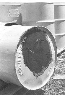 A corroded 55-gallon drum, tipped on its side so the bottom is showing. Grass is growing through holes in the bottom of the barrel.