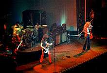 A colour photograph of the four members of Led Zeppelin performing onstage, with some other figures visible in the background