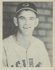 A man smiles for the camera while wearing a dark cap with a "C" on the center and a white baseball jersey.