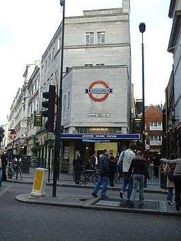 A grey-bricked building with a dark blue, rectangular sign reading "LEICESTER SQUARE STATION" in white letters all under a bright sky