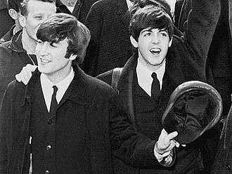 Black-and-white photograph of John Lennon and Paul McCartney in 1964. They are both smiling and waving.