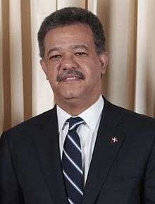 A portrait shot of a middle-aged man smiling somewhat and looking straight ahead. He has light brown skin, slightly African/Negroid facial features, curly dark hair. He is mustachioed and wears a suit and tie.