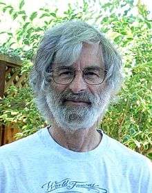 Portrait of a Caucasian man in his seventies with medium-length gray hair and a full gray beard, wearing glasses and a T-shirt.