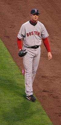 A man in a grey baseball jersey that says "BOSTON" across the chest wearing a red long sleeve shirt underneath the jersey walks on the edge of grass and dirt.