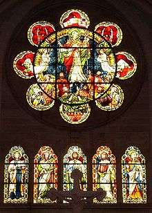 A rose window of simple form having a circle with eight lobes like a flower. It has glass showing Christ rising into heaven watched by apostles and angels. Below are five arched windows each with a saint. Red and yellow are the predominant colours