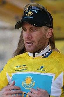A bearded man in his mid-thirties stands in front of a partly visible woman who is zipping up the back of his yellow cycling jersey. He wears a black baseball cap with sunglasses on the cap.