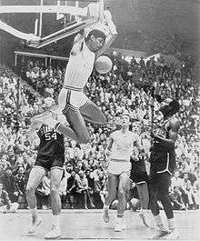 A young black man is completing a two-handed reverse slam dunk during a college basketball game. The photograph is in black and white.