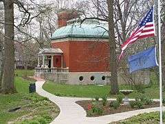 Small red brick building with a green domed roof and flags of the US and Indiana in the foreground