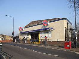 A white-bricked building with a dark blue, rectangular sign reading "LEYTON STATION" in white letters all under a blue sky fading to purple on the horizon