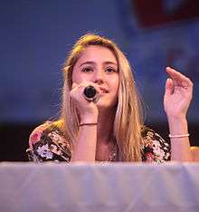 A teenage blonde girl with a black floral patterned top, seated behind a table draped in a white cloth looking forward at an unseen audience; a microphone in her right hand is held to her mouth as she gestures with her left hand.