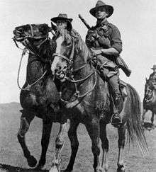 Black and white photo of a man wearing military uniform with a rifle slung across his back riding a horse. Two other similarly-dressed men are partly visible in the background.