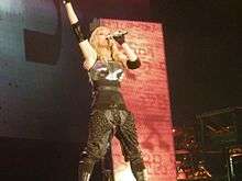 A blond woman singing onstage into a microphone held in her left hand. Her right hand is pointed upwards. The woman is wearing a breast plate and gloves, with knee high boots and ankle covers. Behind her, a rectangular screen displays different symbols.