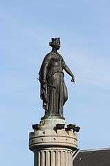 Photo of a statue of a crowned woman wearing a classic Grecian gown. She holds a scepter in her right hand and points down with her left.