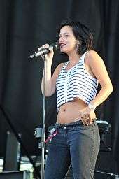 A woman with dark hair, wearing jeans and a cropped white top with black stripes, singing into a microphone