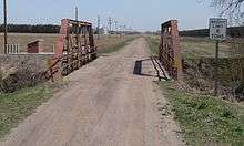 Bridge on dirt road with fields in distance; railcars at right edge of picture
