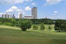 A green space with trees and rolling lawns is flanked by tall, modern-style buildings in the background on a sunny day.