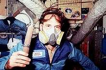 A man holding a piece of hose floats in front of a selection of transient space station hardware. He is wearing a gray-and-yellow plastic mask over his mouth and nose, a pair of goggles above his eyes, and a blue jumpsuit with a name patch on it.