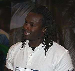 A black man with dreadlocks wearing a white polo shirt. He is smiling and facing left.